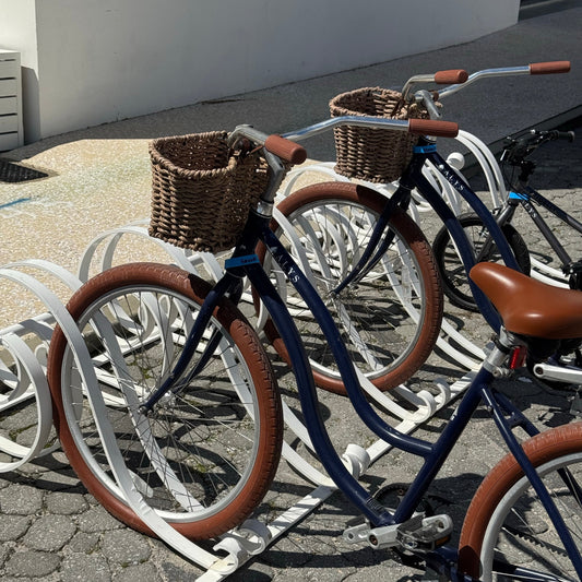 Two bicycles with brown baskets on a bike rack against a white wall.